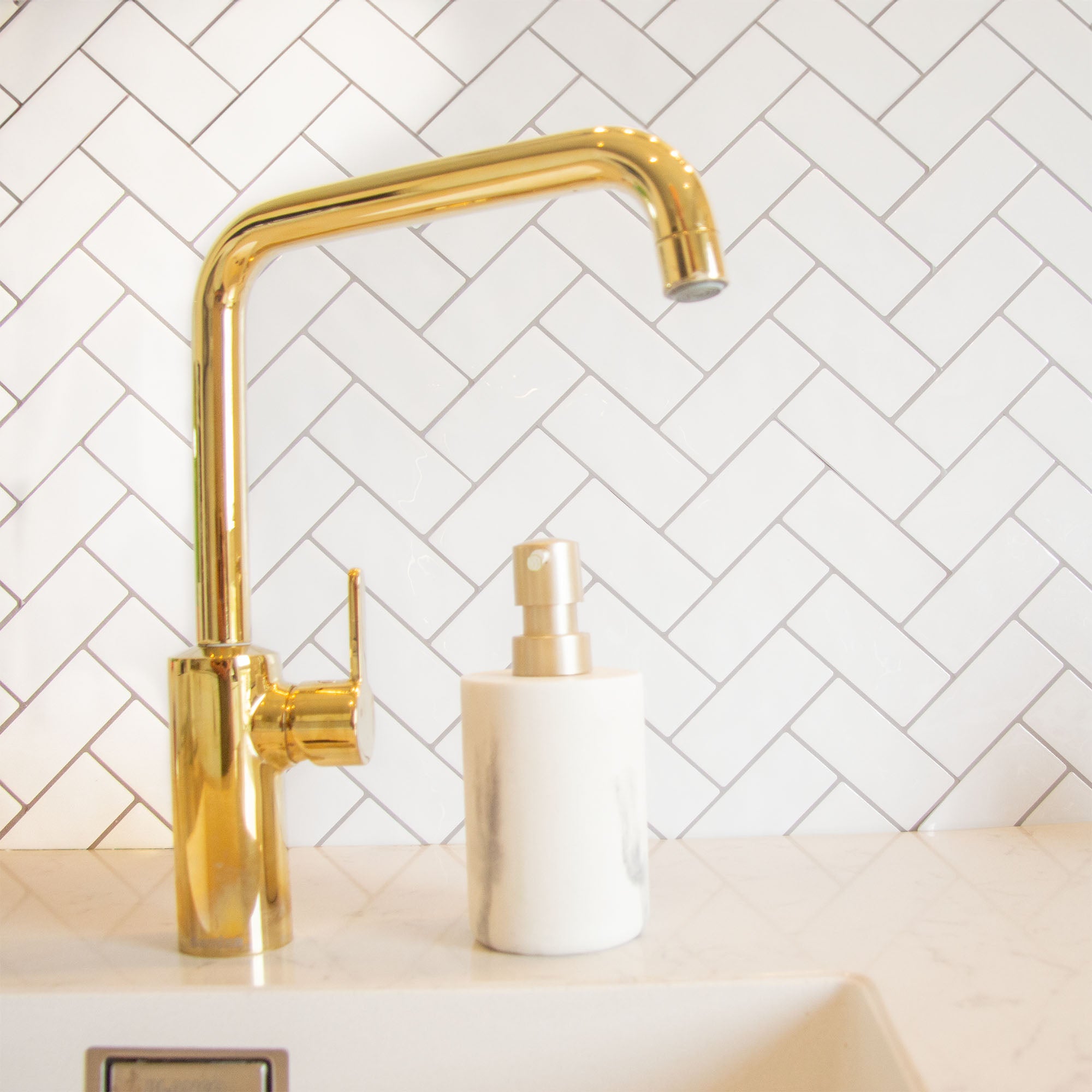 Hvid peel and stick flise (snowbone) fra stickoramic i sildebensmønster. Brugt som backsplash i køkken med hvid håndvask og guld vandhane.